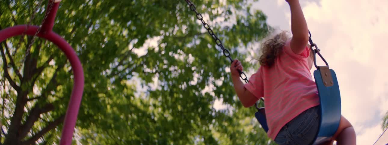 A low-angle, slow-motion view of two carefree young girls swinging at a park on a bright summer day