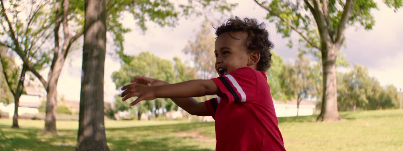 Slow motion shot of a joyful toddler with curly hair laughing and playing with bubbles in a green park