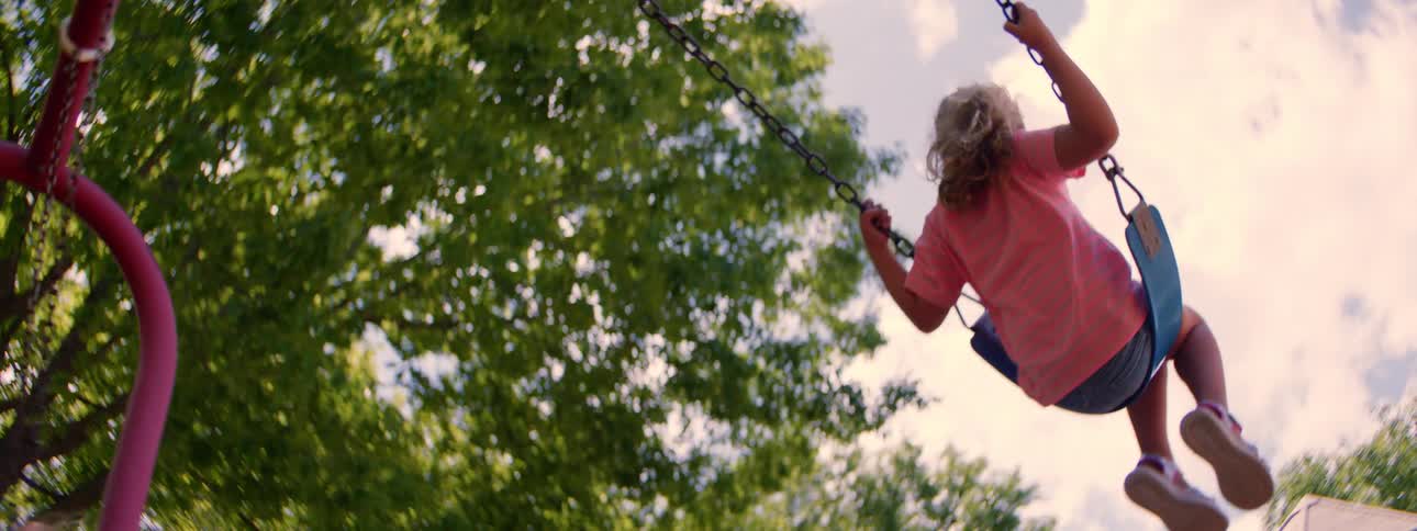 Low angle slow motion shot of a young girl swinging towards the sky under lush green trees
