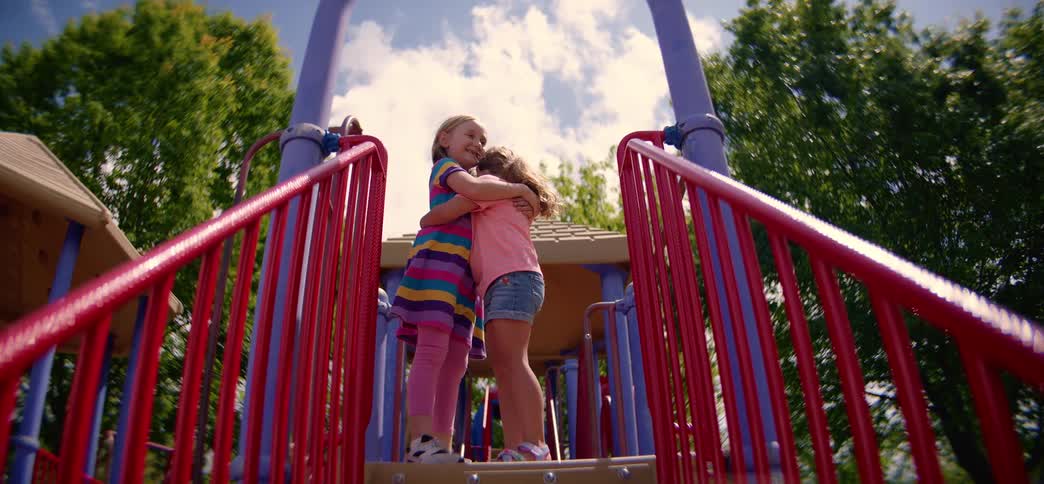 Two smiling little girls share a long, affectionate hug on a playground structure on a bright, sunny day