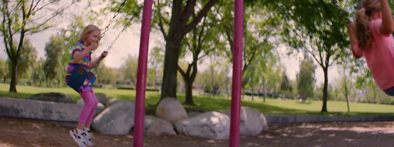 Two young girls smile and laugh as they swing back and forth in slow motion at a park on a sunny day