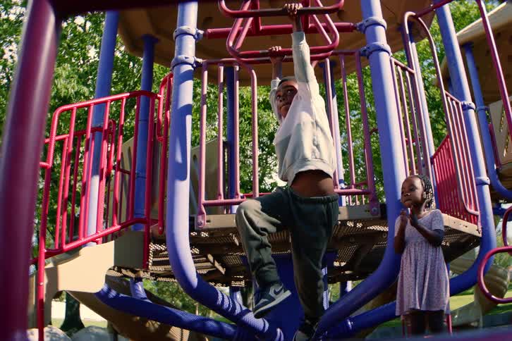 A young boy plays on the monkey bars at a sunny park playground while his sister watches and claps in encouragement