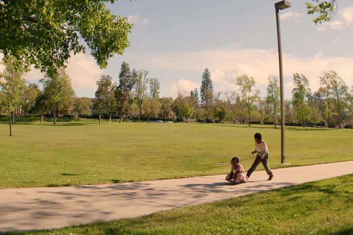 A little boy pushes his younger sister on a skateboard along a path in a sun-drenched park in slow motion