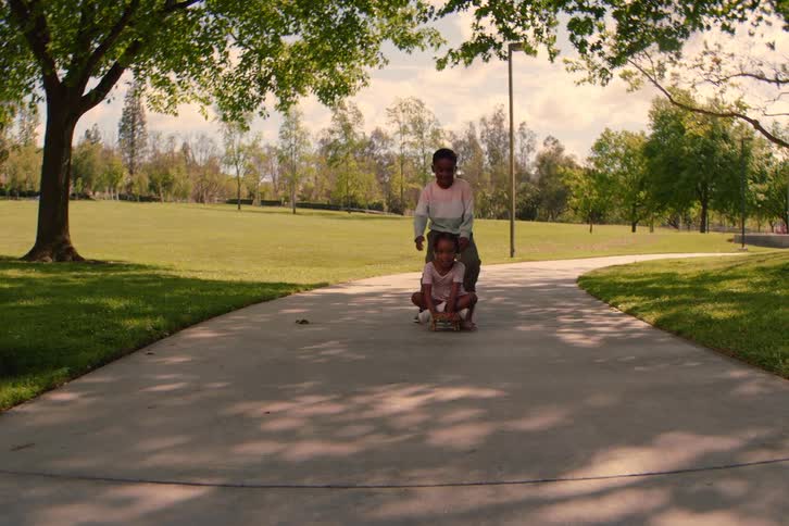 A young boy pushes his little sister on a skateboard as they ride towards the camera in slow motion through a sunny park