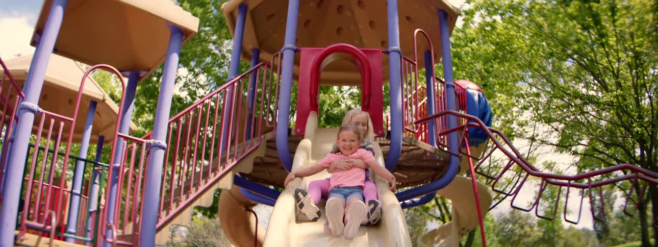Two happy sisters smile and laugh as they go down a playground slide together in slow motion on a sunny day
