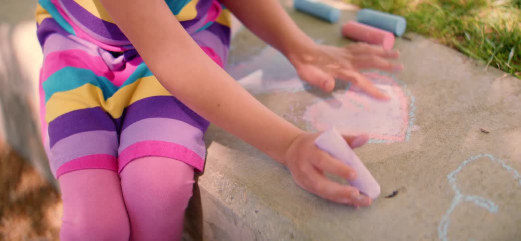 A young girl sits outside and uses her hands to blend sidewalk chalk inside a heart she drew on the concrete