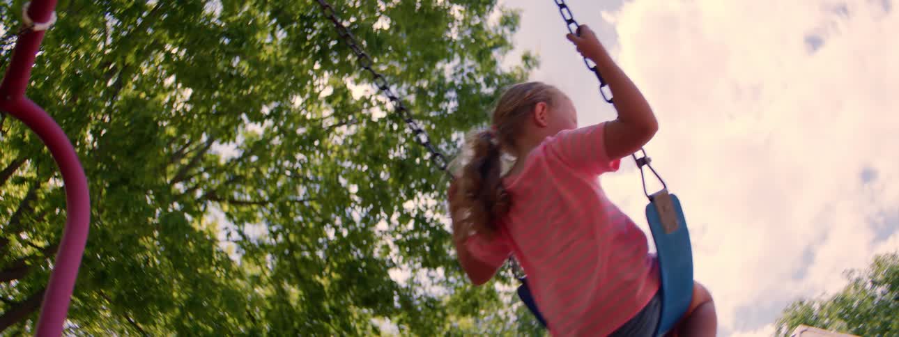 Low angle slow motion shot of a young girl with a ponytail swinging on a swing set under leafy green trees