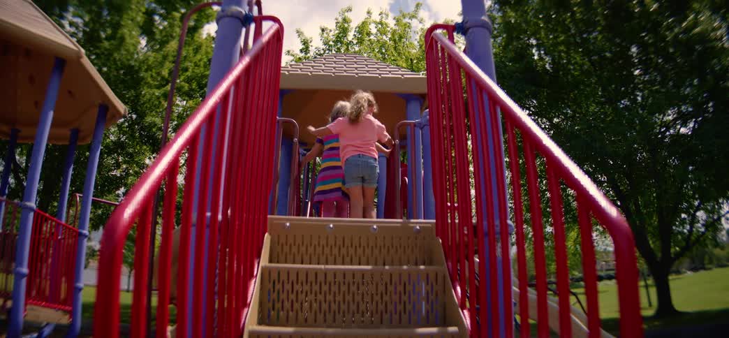 Two young girls happily race up the stairs of a colorful playground structure in a sunny park