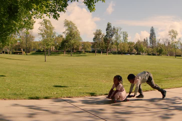 A big brother pushes his little sister on a skateboard down a sidewalk path at the park in super slow motion