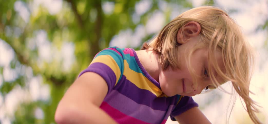 A close-up of a young blonde girl in a colorful striped shirt concentrating on an activity outside on a sunny day