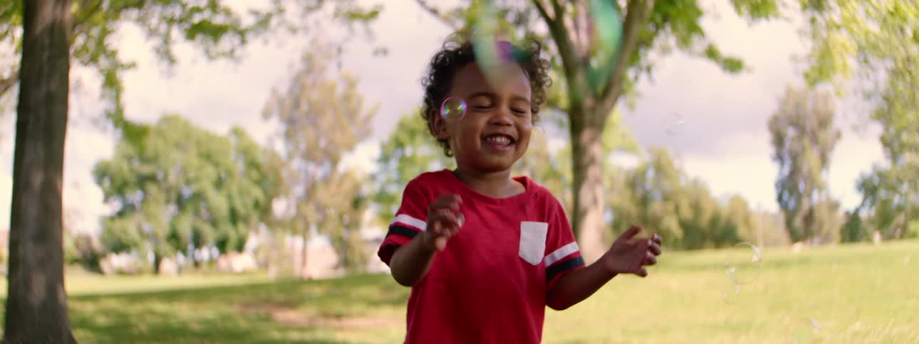 Slow motion of a happy young toddler with curly hair laughing and playing with soap bubbles in a park on a sunny day