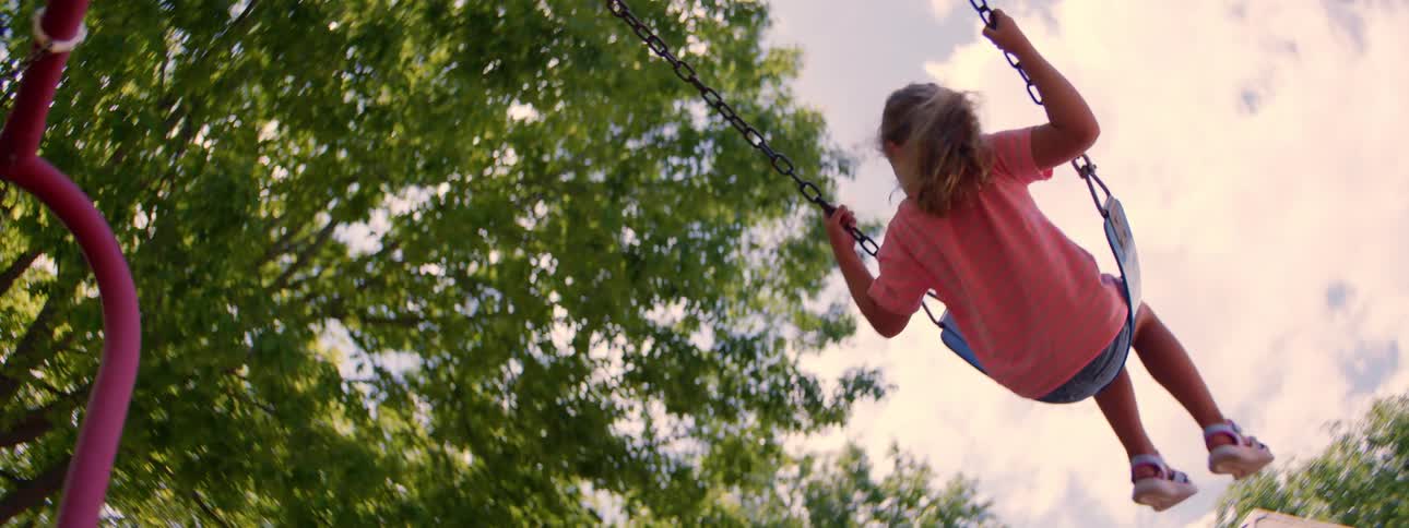 Low angle slow motion shot of a little girl with blonde hair swinging back and forth on a swing set at a park on a sunny day