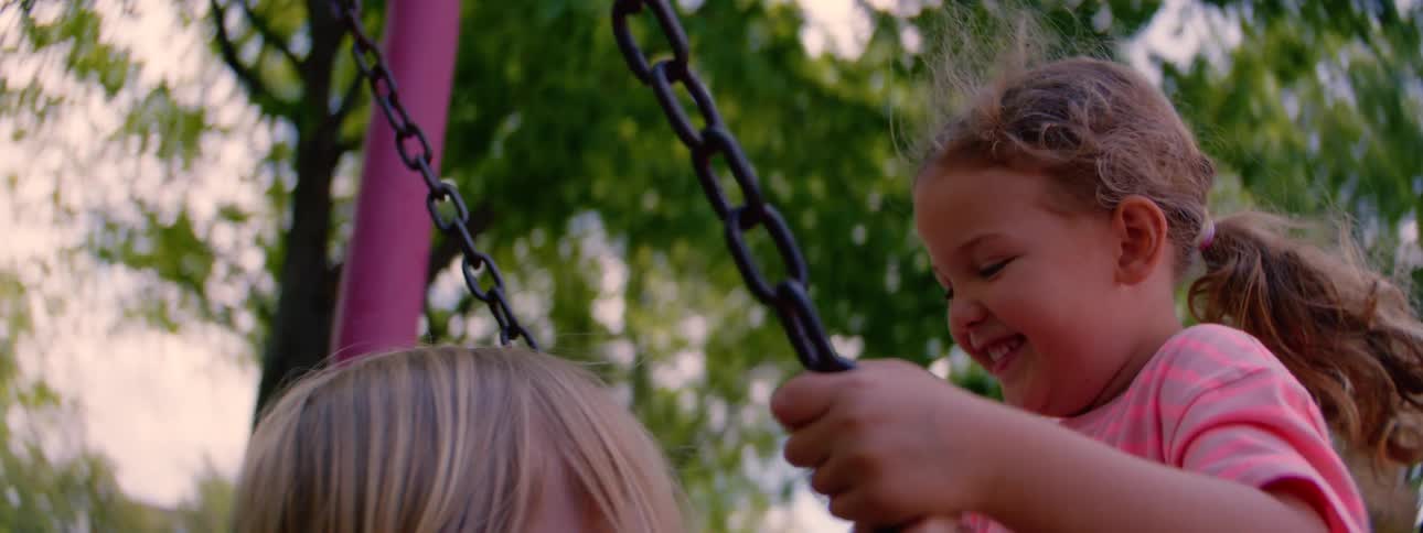 Two young girls smile and laugh as they swing together at the park in slow motion