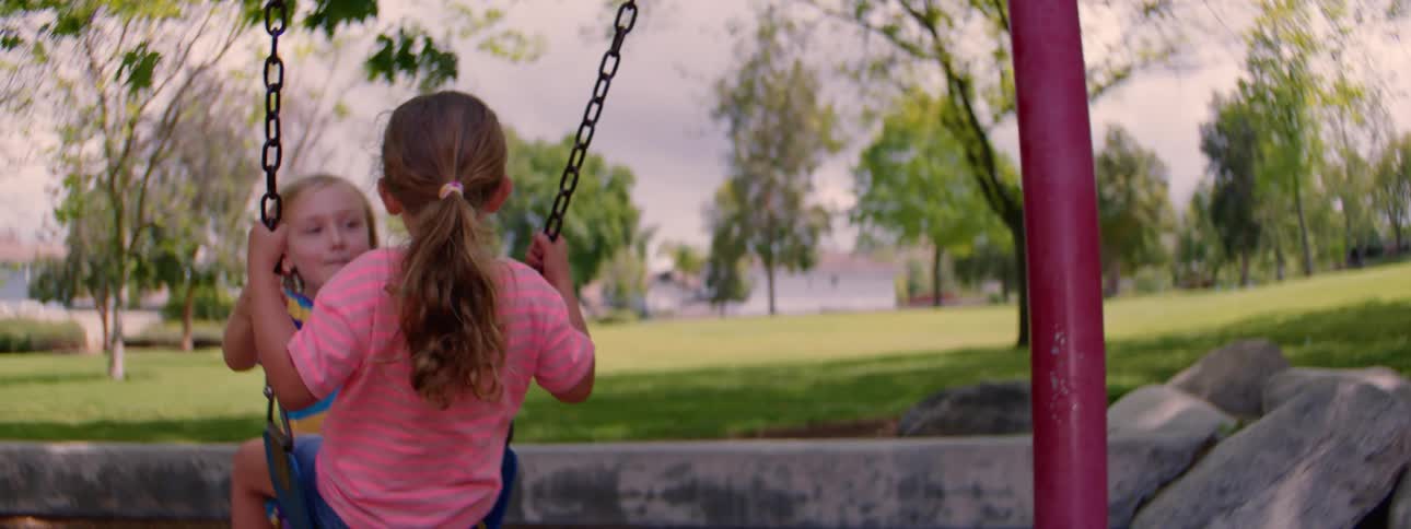 Two young friends swing together on a swing set at a park on a sunny day in slow motion