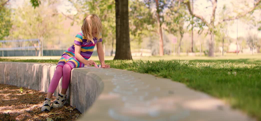 A little girl in a colorful striped shirt draws with chalk on a concrete wall in a sunny park
