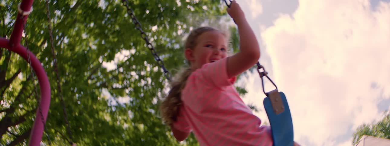 A happy little girl with blonde hair smiles as she swings in slow motion on a sunny summer day, filmed from a low angle
