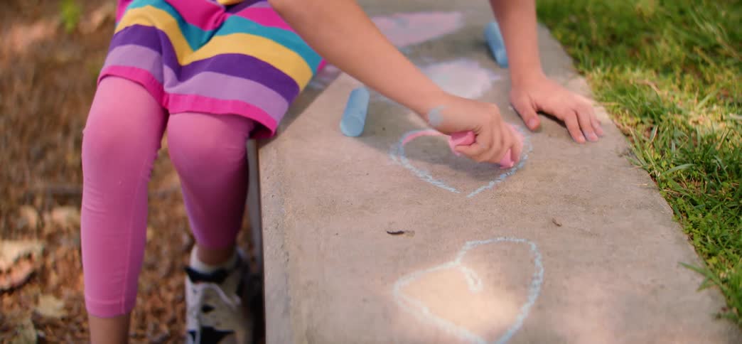 Close up on a little girl's hands as she uses pink chalk to color in a heart drawing on a concrete step outdoors