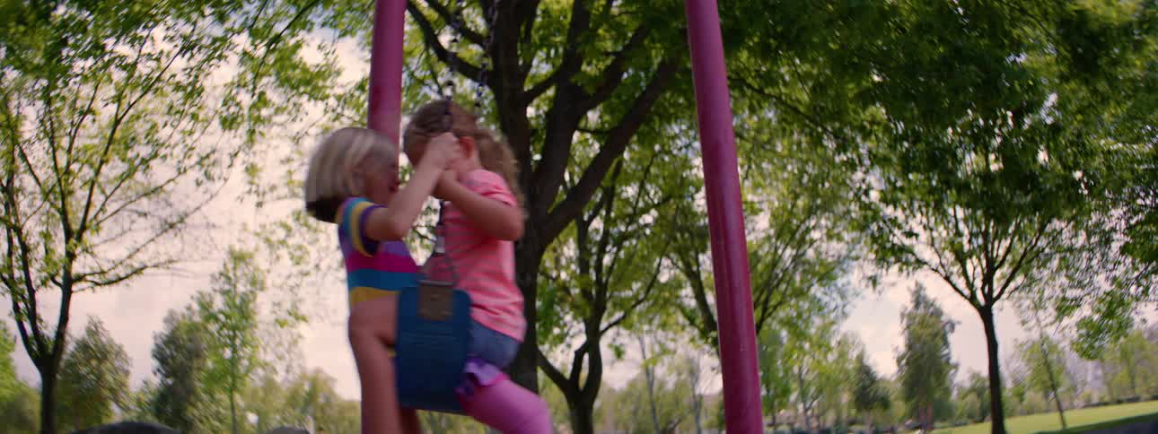 A joyful slow-motion shot shows two little girls sharing a swing and laughing at the park