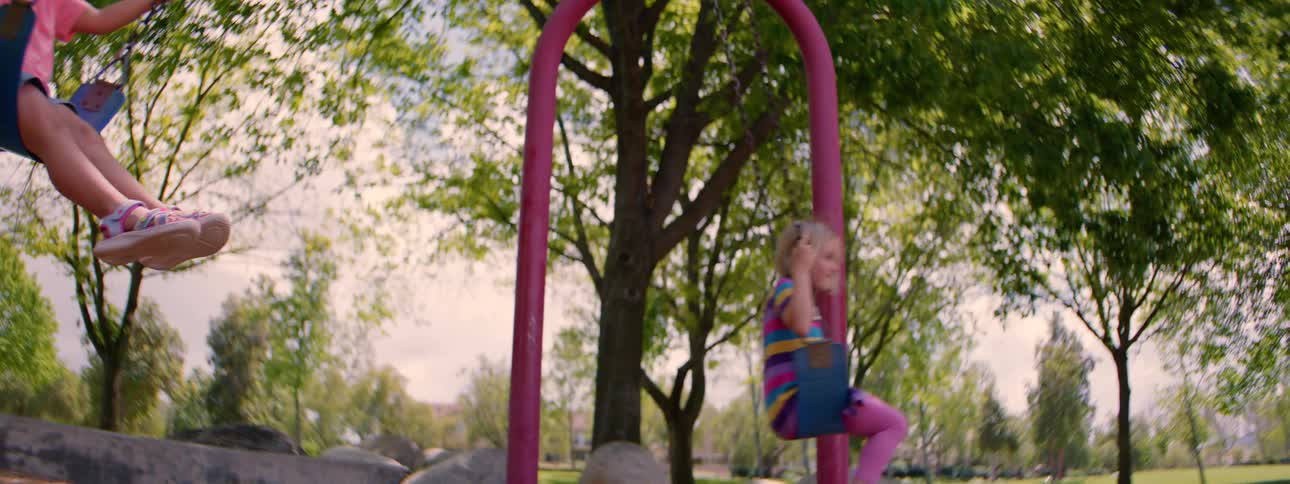 A low-angle slow-motion shot captures two happy young girls swinging back and forth at the park