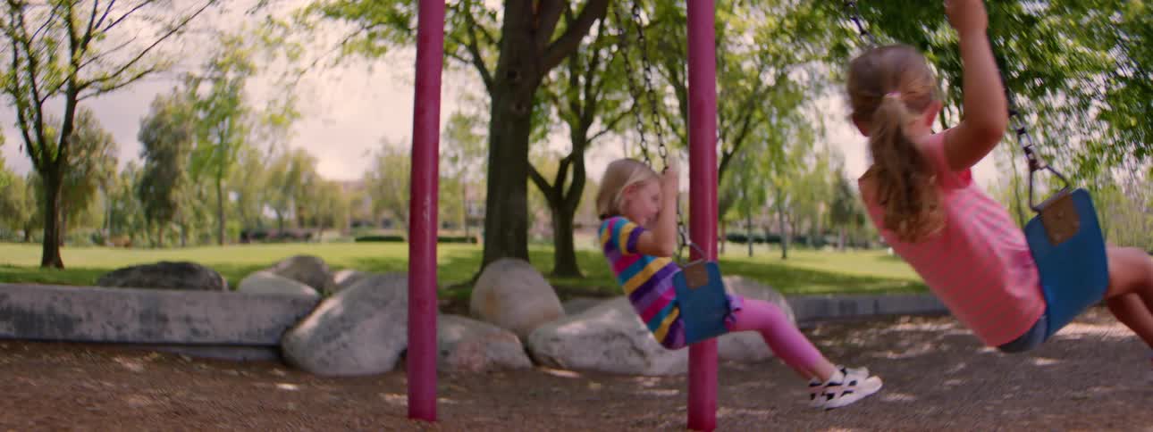 Two young sisters swing back and forth in slow motion on a playground swing set
