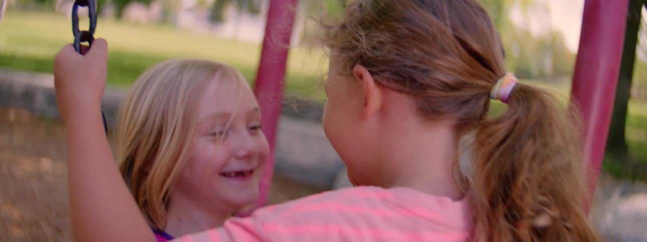 A slow-motion close-up of two young friends smiling and playing together on a park swing on a sunny day