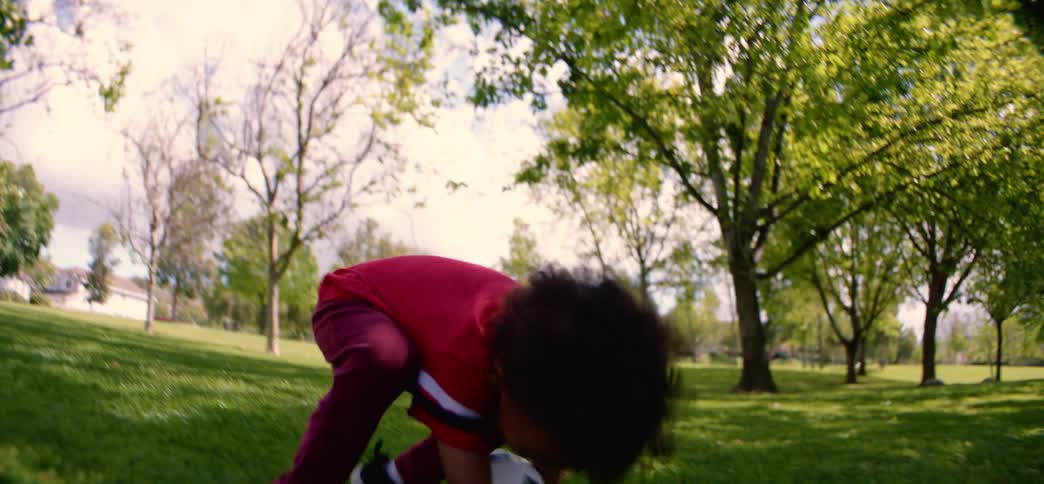 An adorable little boy with curly hair laughs as he throws and chases a soccer ball across a grassy field in slow motion