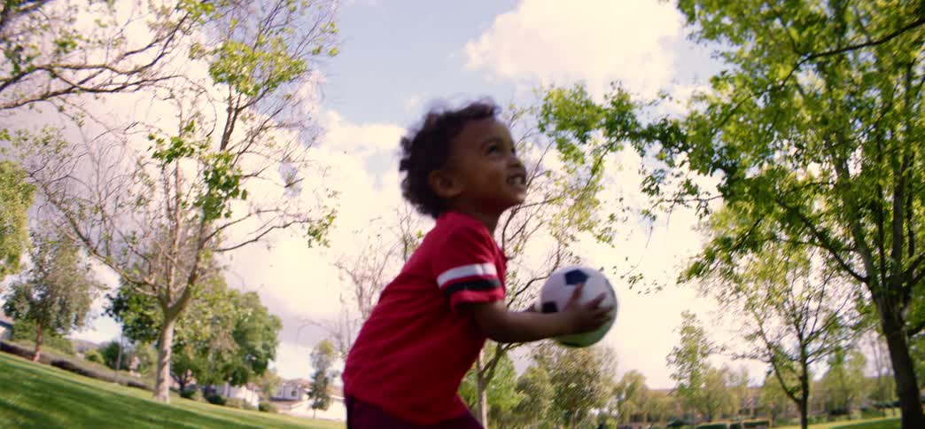 A gleeful toddler enjoys a sunny day in the park, playing with his soccer ball in slow motion