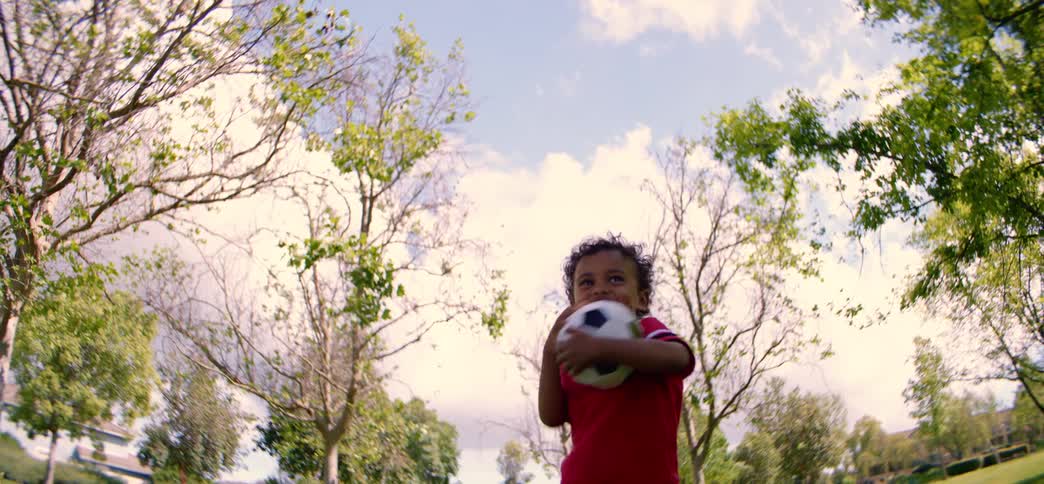 Slow motion, low angle shot of a happy toddler running with a soccer ball in a park on a sunny day