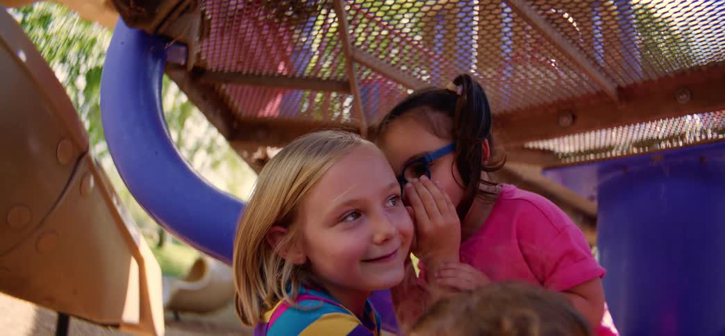 Close up of two adorable little girls sharing secrets and smiling while playing on playground equipment on a sunny day