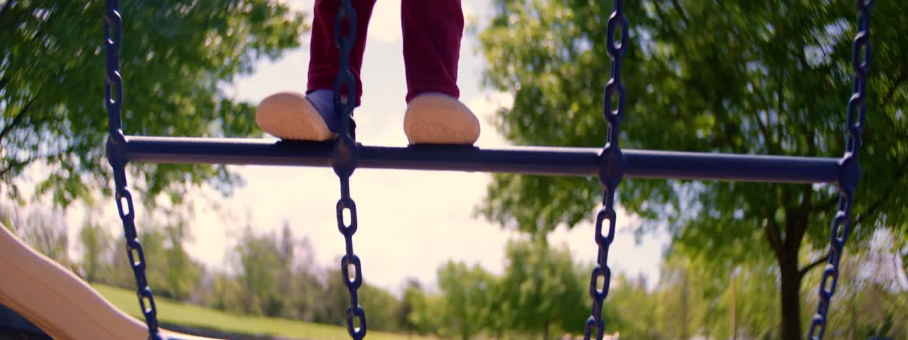 A low angle close-up shows a child's feet in blue shoes carefully balancing on a playground structure on a sunny day