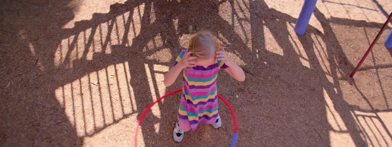A high-angle slow-motion shot captures a little girl in a colorful striped dress learning to hula hoop at the park