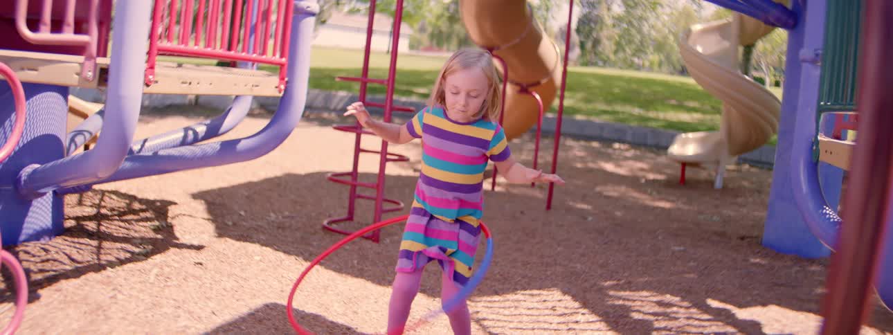 Slow motion shot of a cute girl in a striped dress trying and failing to hula hoop at a playground