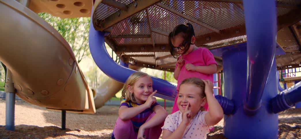 Three happy little girls share a secret and play together under the playground equipment on a sunny day