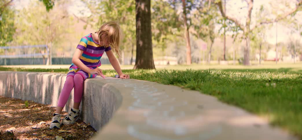 A creative little girl sits on a concrete ledge in a sunny park and draws hearts with chalk