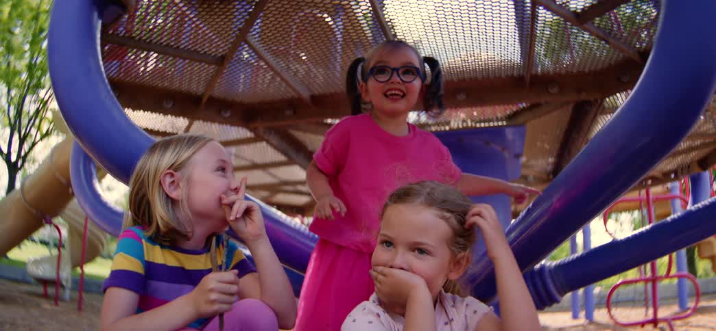 Three adorable little girls smile and laugh while sharing secrets on a colorful playground on a sunny day