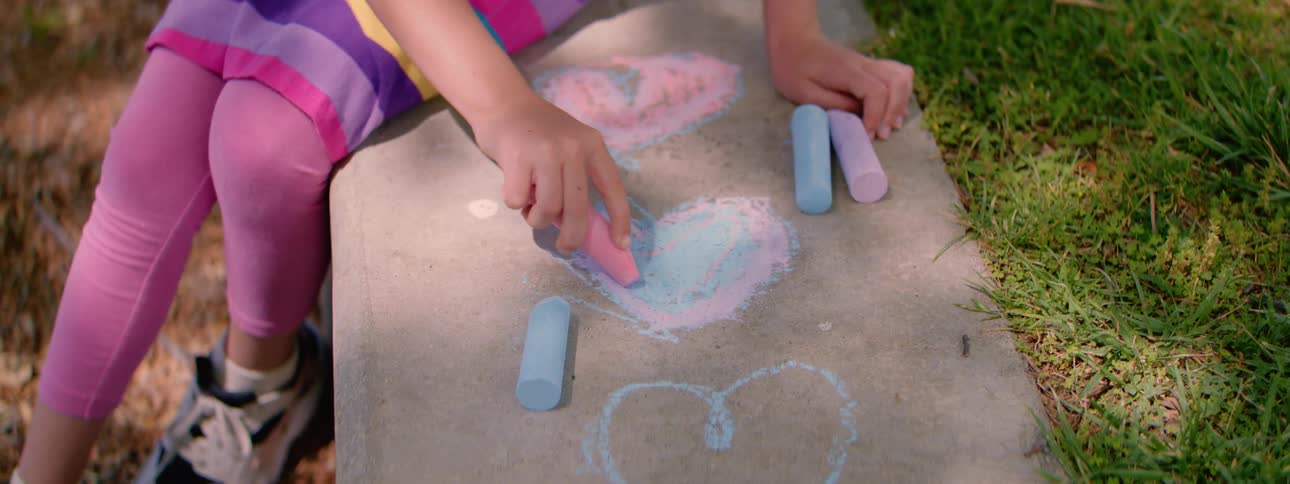 A young girl colors in a heart shape with pink and blue chalk on a sidewalk on a sunny day