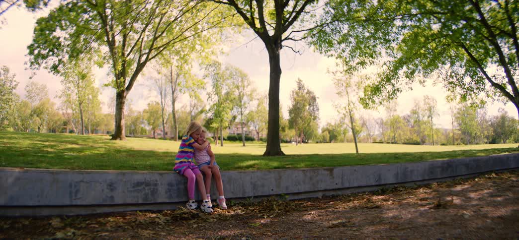 Adorable little sisters share a sweet hug while sitting together on a wall in a beautiful, sunny park