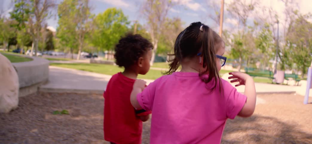 Two toddler friends, a young boy and a girl with pigtails, walk through a sunny park together in slow motion