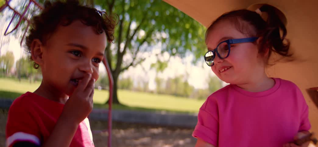 Two adorable toddlers, a boy and a girl, laugh and play together in slow motion on a sunny playground