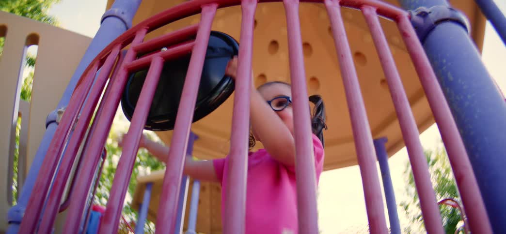 An adorable little girl with pigtails and glasses joyfully plays on a colorful playground, smiling and laughing from behind the bars