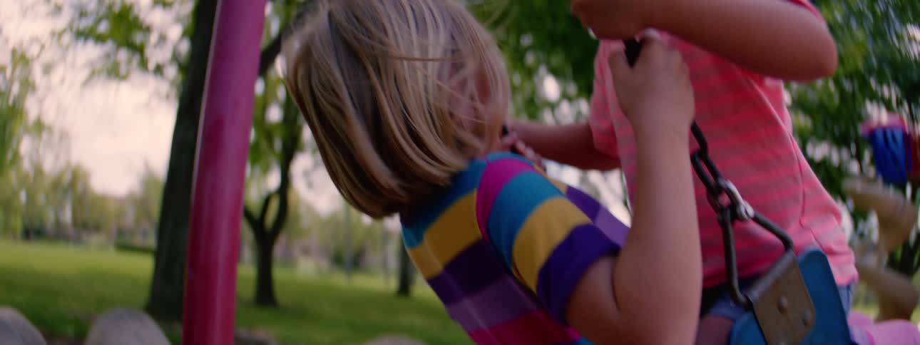 A slow motion, tilt-up shot reveals two young girls happily swinging together on a swing set at the park