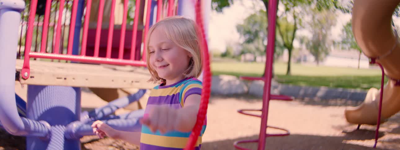 Slow motion shot of a smiling little girl with blonde hair happily spinning a hula hoop on a sunny day at the park playground