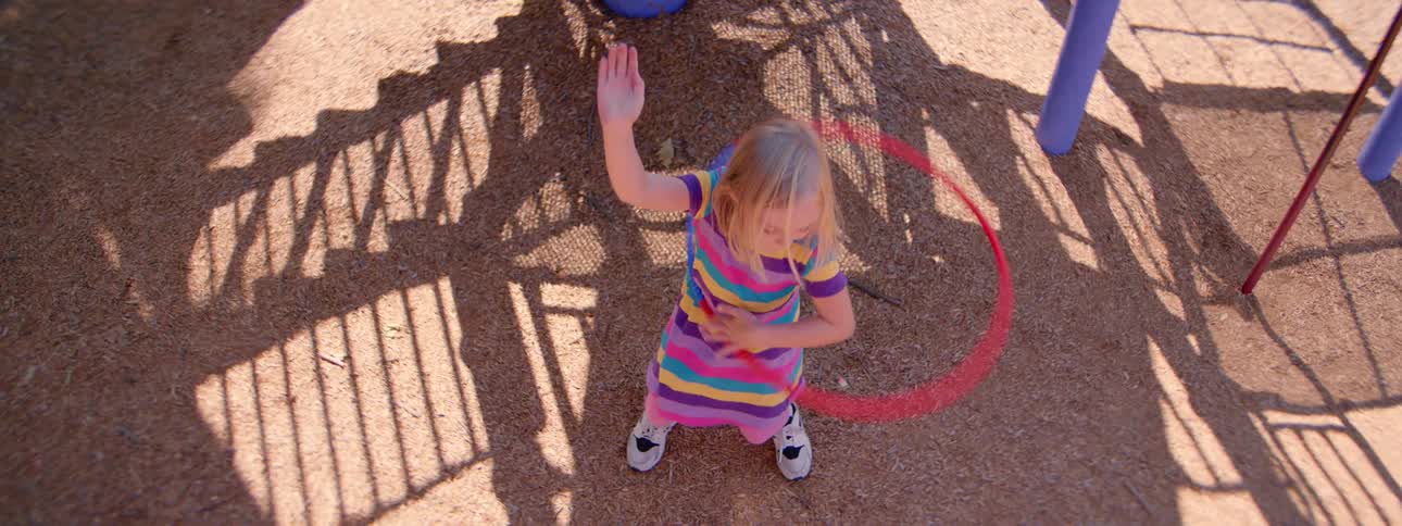 A young girl in a colorful striped dress attempts to hula hoop in slow motion at the playground, as seen from an overhead view