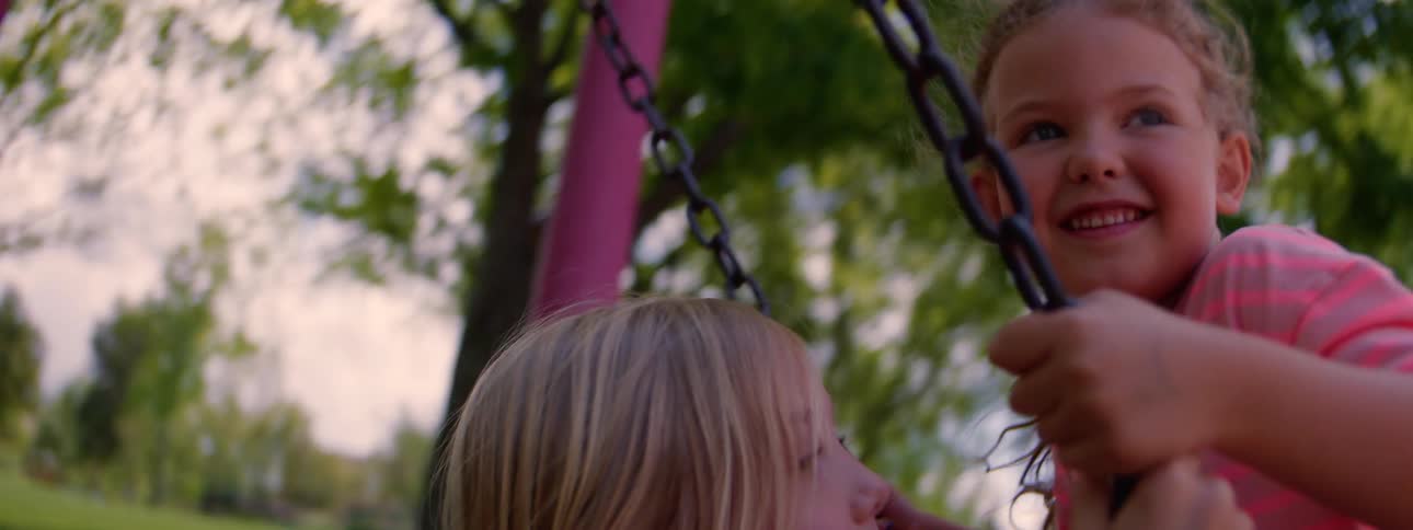 Two happy young girls smile at each other while sharing a swing at the park in slow motion