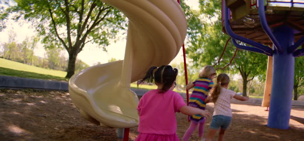 Joyful young girls play together on a sunny day at the park playground, with one sliding down a slide in slow motion