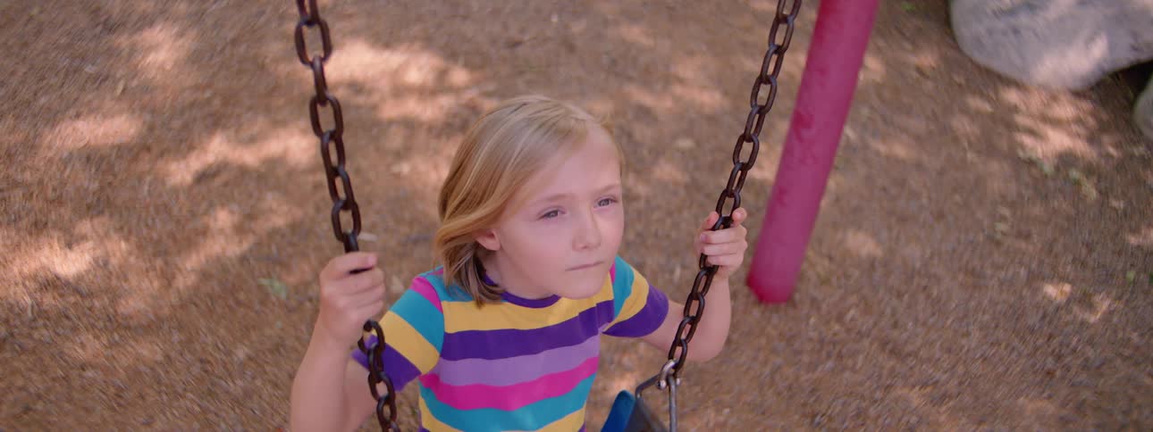 A sad and lonely young girl sits on a swing at the playground looking thoughtful