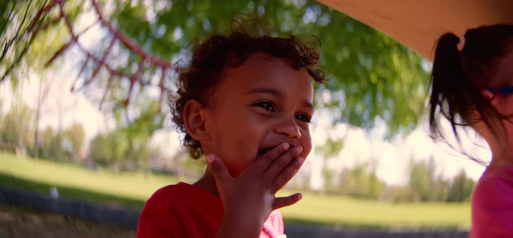 An adorable toddler girl shares a berry with her friend, eliciting a joyful laugh from him in slow motion at the park