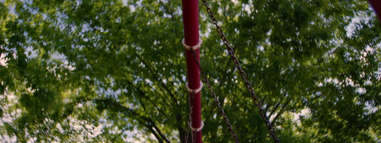 An upward-looking view shows a red swing set pole and its chains moving gently, with green trees out of focus in the background