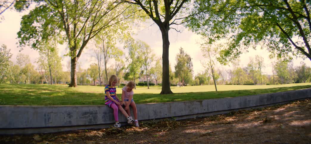 Two little girls sit together on a concrete wall at the edge of a lush green park on a bright, sunny day