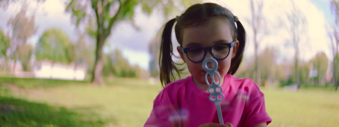 Close up of a cute young girl with pigtails and glasses blowing bubbles in a park in slow motion